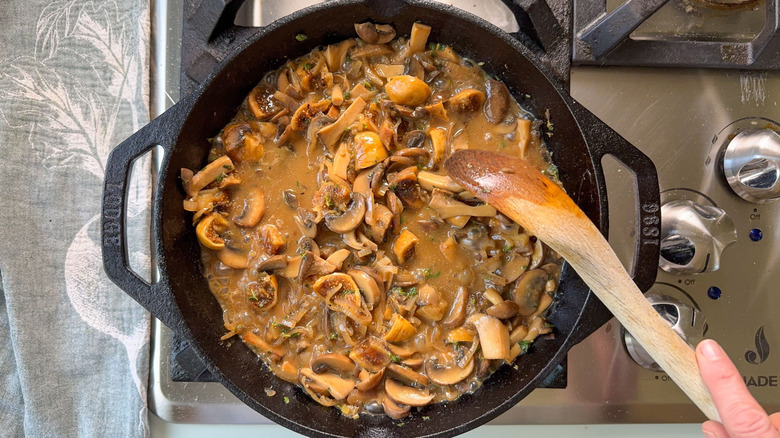 Stirring aromatic smothered pork chops sauce in cast iron skillet on stovetop with wooden spoon