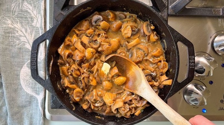 Stirring butter into aromatic smothered pork chops sauce with wooden spoon in cast iron skillet on stovetop
