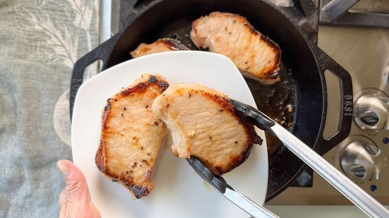 Removing browned pork chops from cast iron skillet to a plate with tongs