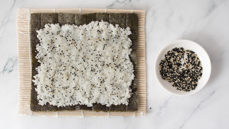 Nori sheet with rice and sesame seeds next to bowl of sesame seeds