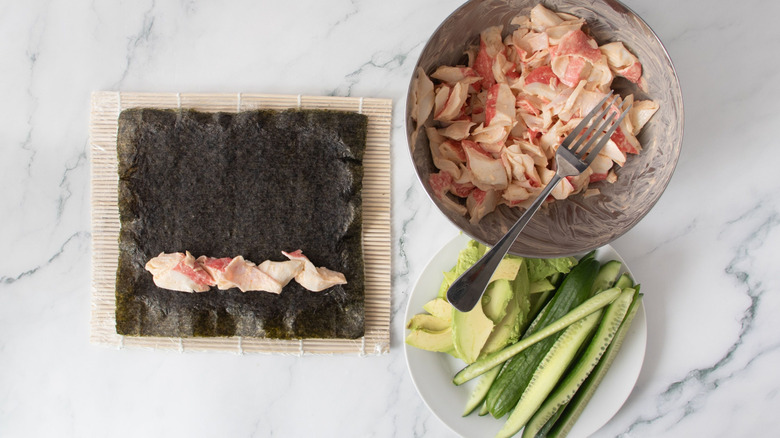 Nori sheet with crab mix next to bowl of crab mix and plate with cucumber and avocado