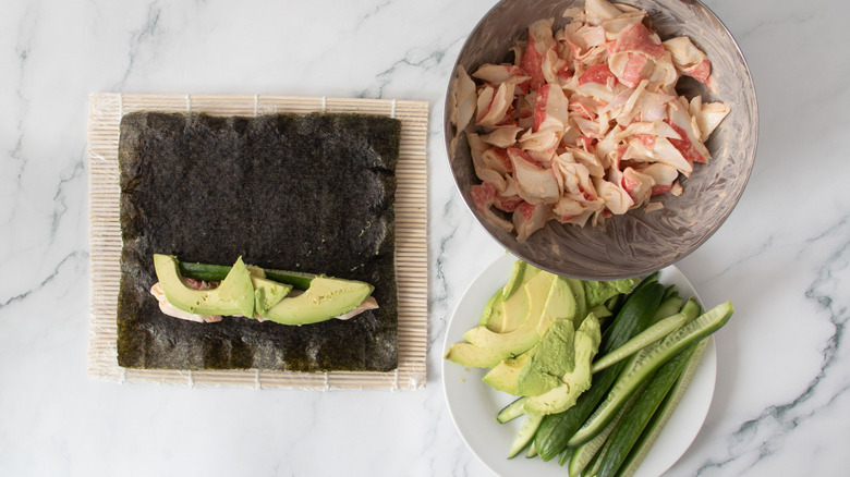 Unrolled California roll next to bowl of crab mix and plate with cucumber and avocado