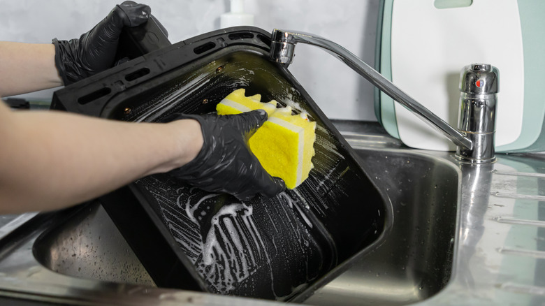 A person cleaning the basket of an air fryer with a sponge and soap.