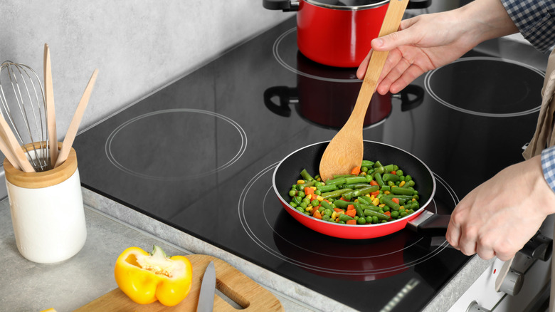 Person stirring vegetables in a pan with a wooden spoon on a glass cooktop electric stove