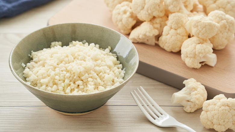 A bowl of cauliflower rice next to pieces of cauliflower on a cutting board and a fork