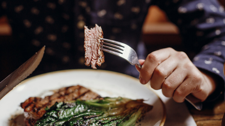 Person holding a fork with a bite of steak on it over a plate of steak with cabbage