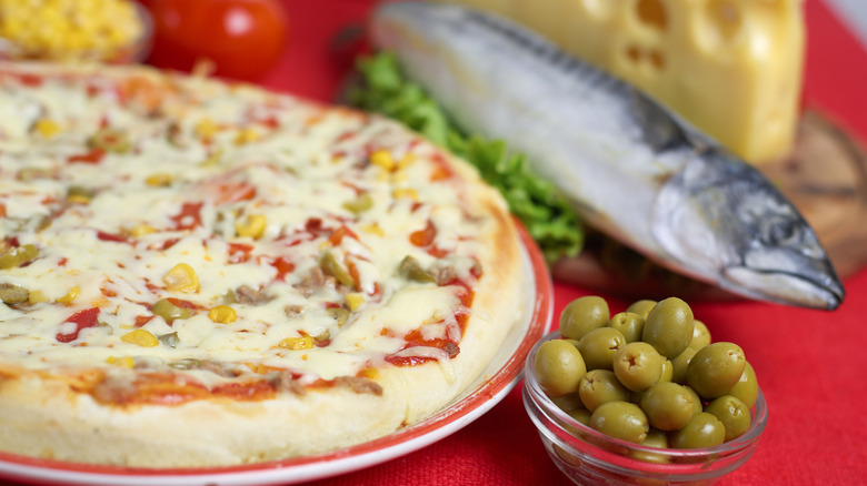 An undercooked pizza next to a whole fish and a bowl of green olives