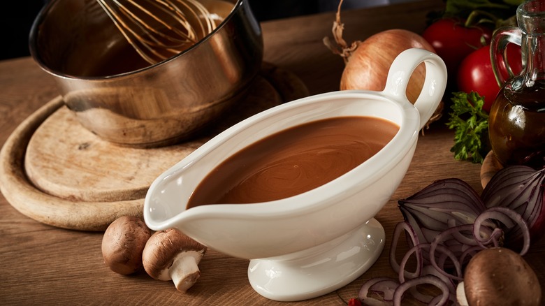 A gravy boat filled with gravy on a wood table with mushrooms, onions, tomatoes, bowl, whisk, cutting board, and bottle of oil