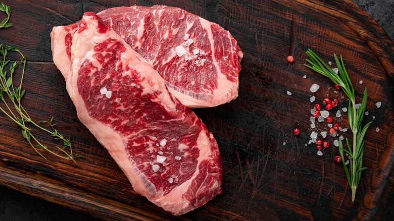 Two raw New York strip steaks with salt, peppercorns, and herbs