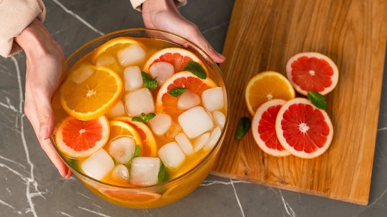 Large punch bowl filled with ice and assorted citrus next to wooden cutting board.
