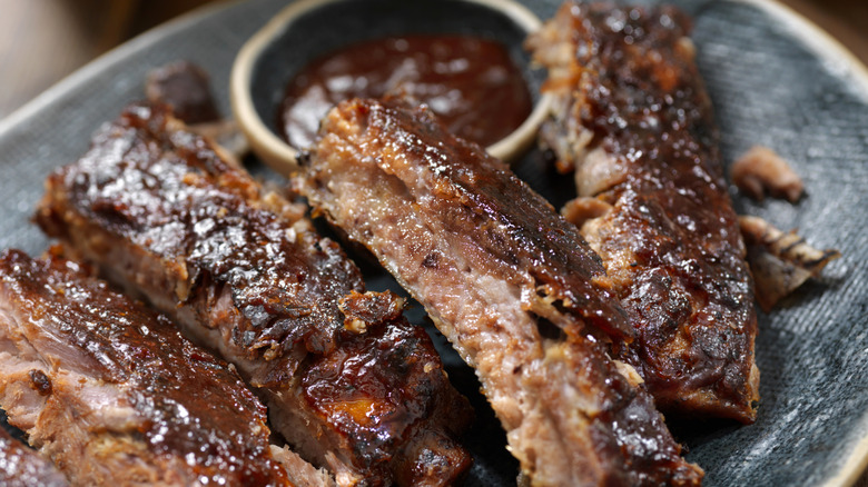 Slow-cooked, St. Louis-style baby back pork ribs slathered in barbecue sauce sit on a plate, alongside a cup of extra sauce.