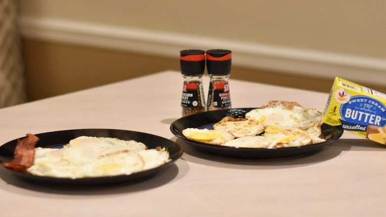 Two plates of fried eggs with bacon, butter, salt, and pepper