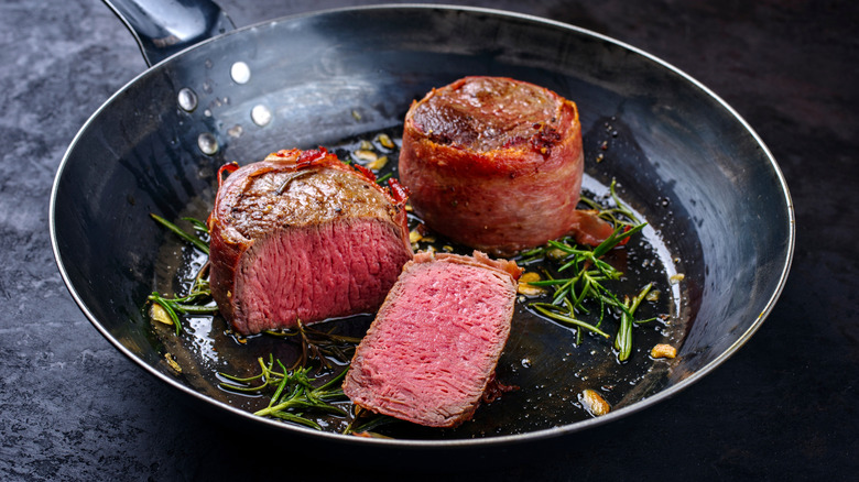 Filets wrapped in bacon cooking in a frying pan with rosemary