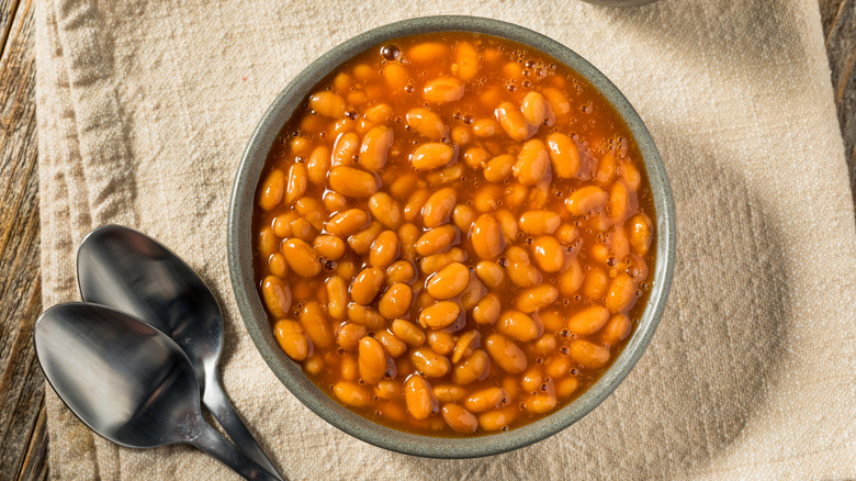 Barbecue Baked Beans with Tomato Sauce in a bowl with two spoons on a fabric tablecloth