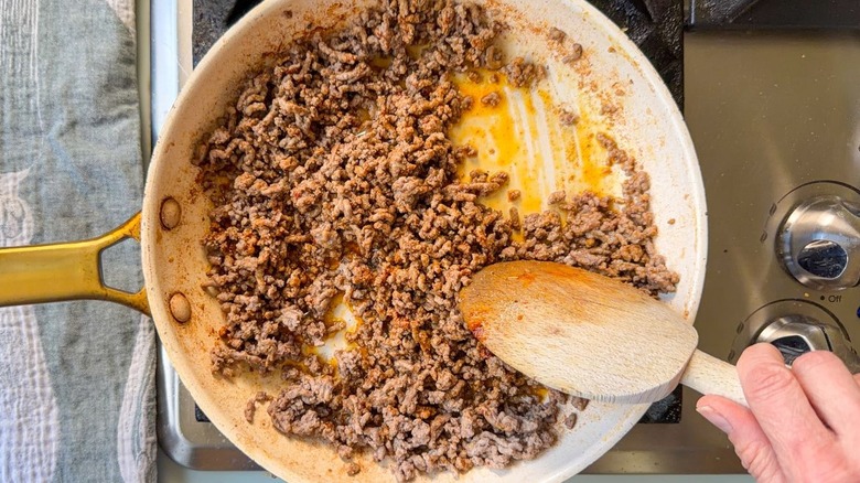 Stirring browned ground beef and spices with wooden spoon in skillet on stovetop
