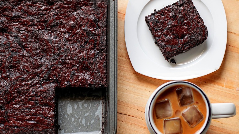 One brownie on a small white plate next to a pan of brownies and a mug of iced coffee.