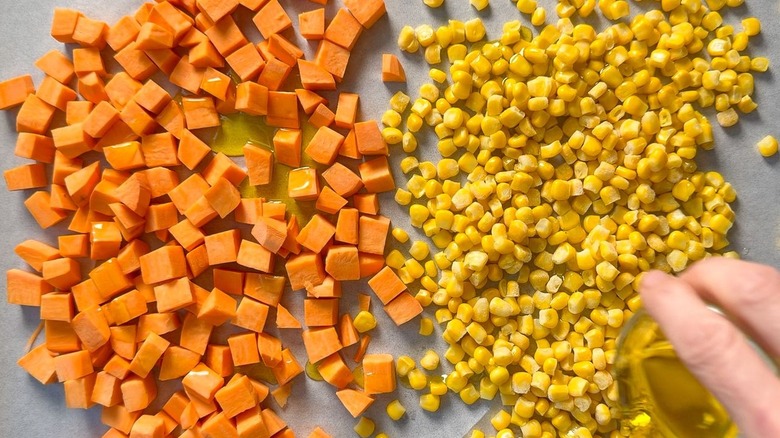 Adding olive oil to cubed sweet potatoes and corn kernels on parchment paper-lined baking sheet