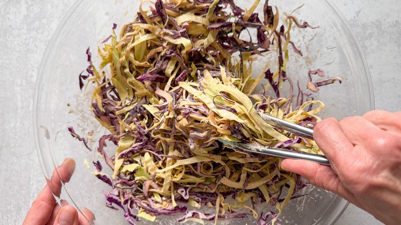 Tossing shredded purple and green cabbage with dressing in glass bowl with tongs