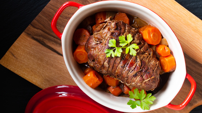 A beef pot roast surrounded by carrot pieces with herbs in deep pot on cutting board.