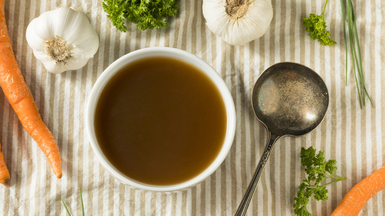 A white bowl of beef broth is photographed from above with garlic and carrots in the frame.