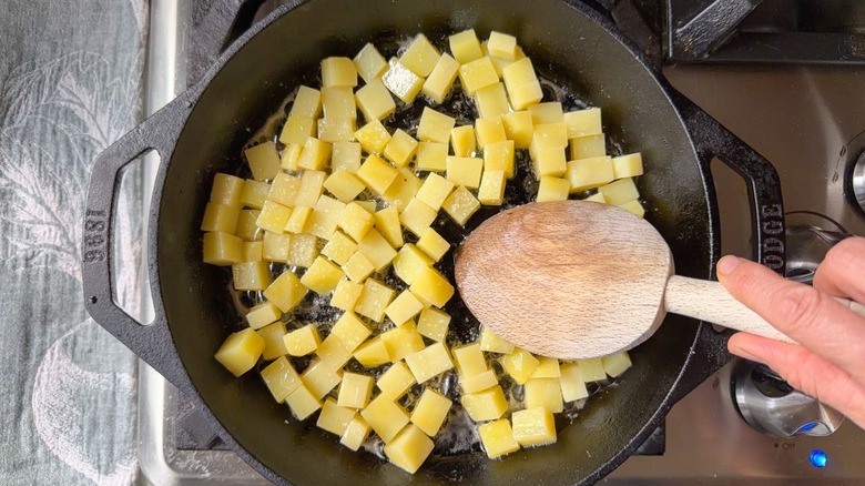 Stirring cubed potatoes in butter in cast iron skillet with wooden spoon on stovetop