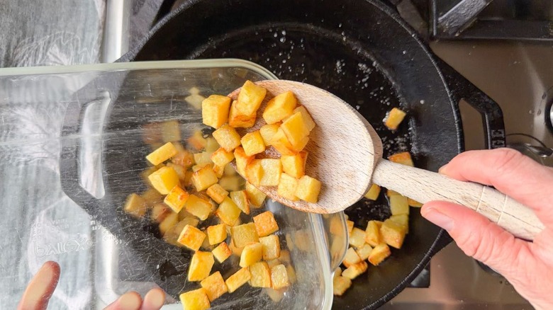 Spooning cooked cubed potatoes into glass baking dish from cast iron skillet on stovetop