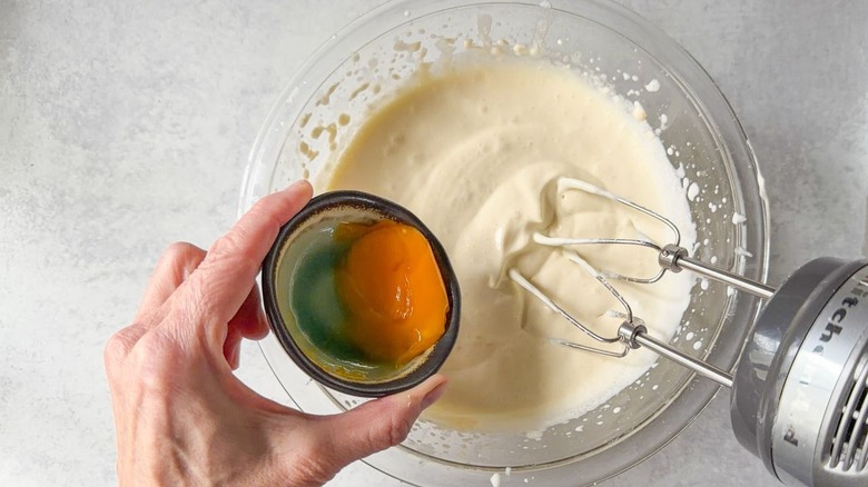Adding an egg yolk to softly beaten heavy cream in glass bowl