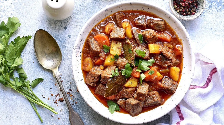 Beef stew in a white-speckled bowl next to a metal spoon and a white cloth napkin with a red stripe