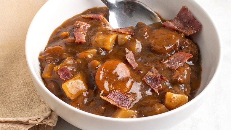Close-up of beef stew with carrots, potatoes, and bacon in a white bowl with a metal spoon