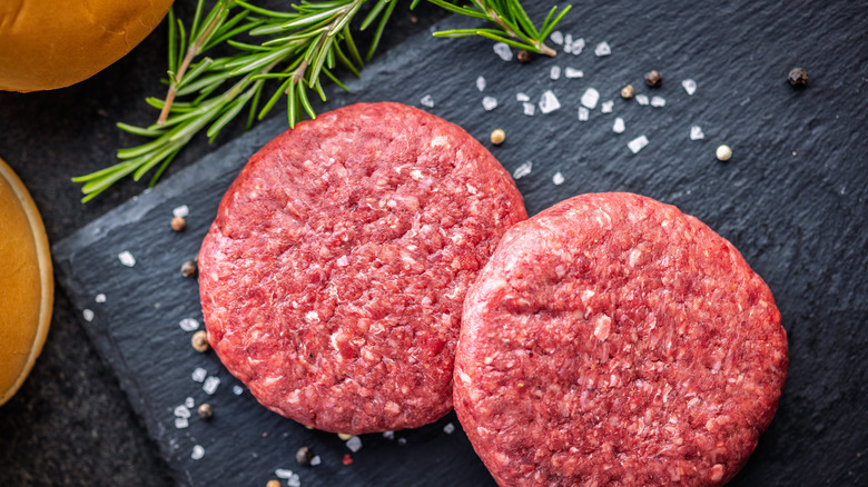 Two uncooked burger patties on a black cutting board.