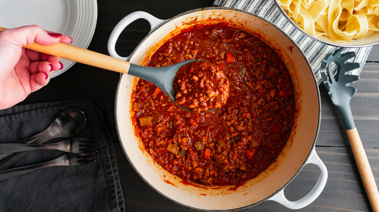 Person preparing Bolognese sauce in Dutch oven, a bowl of pasta visible in background