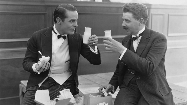 Two men eating sandwiches and drinking milk outdoors, early 1900s