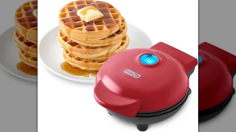 Small red Dash waffle maker with an illuminated blue light on top next to a stack of waffles on a white plate with syrup and butter