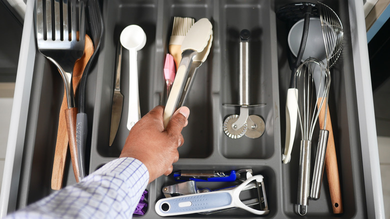 Person's hand placing tongs into a gray organizer in a kitchen drawer filled with other kitchen tools