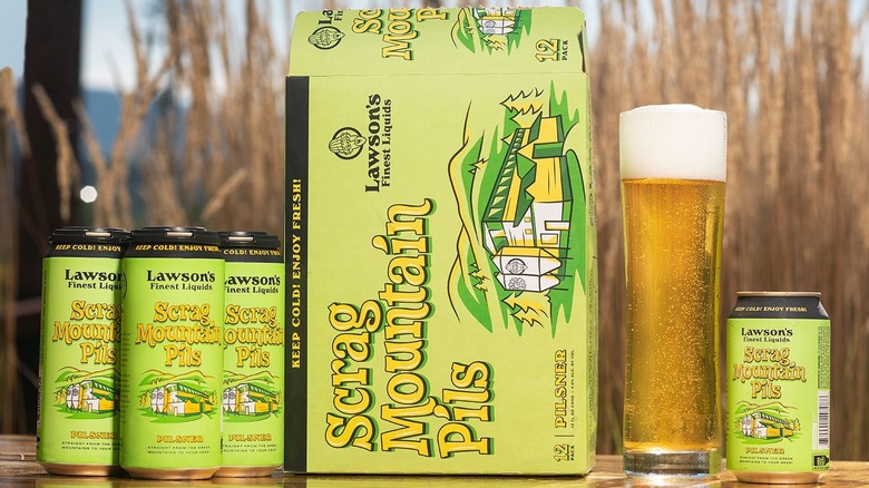 A case, cans, and glass of Lawson's Finest Liquids Scrag Mountain Pils lined-up on an outdoor tabletop
