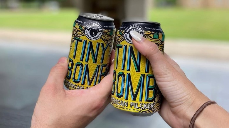 Two hands toasting cans of Wiseacre Tiny Bomb American Pilsner