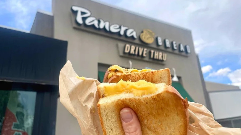 A person's hand holding a grilled cheese sandwich in front of a Panera Bread location