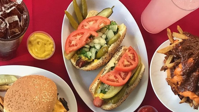 A pair of Chicago style hot dogs on a white plate with an assortment of other food on a red table