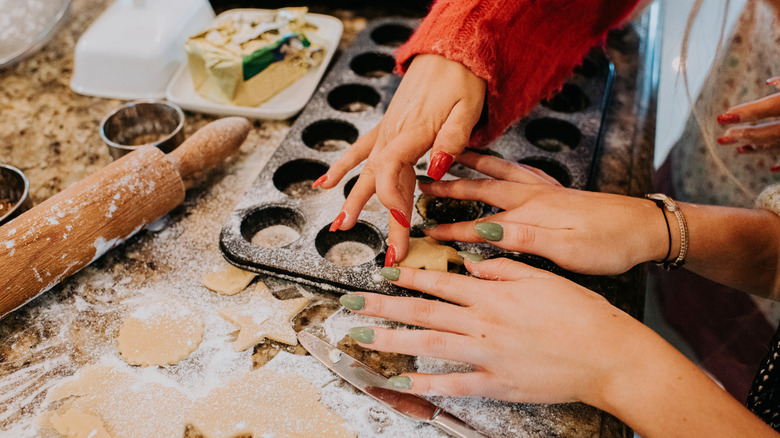 A close-up of two people shaping and cutting out dough for Christmas cookies, one with red painted nails and the other with green painted nails