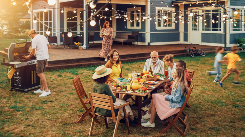 Group of people gathered around a table at a backyard BBQ while a man is grilling, a woman is walking over, and kids are running around