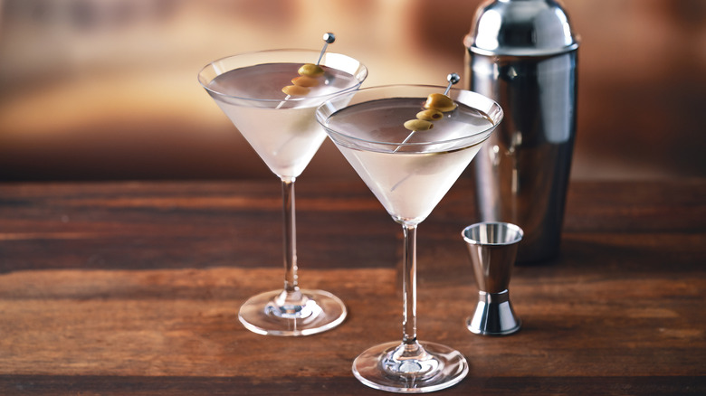 Two glasses of dry martini with olive sit on a wooden bar next to a cocktail shaker