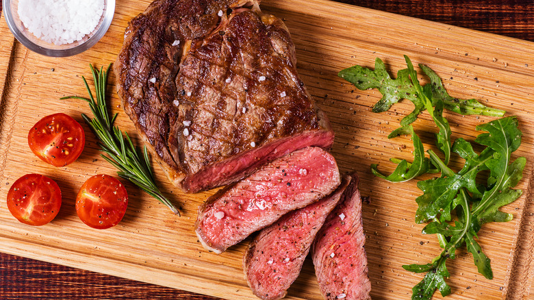 A medium-rare, partially cut ribeye steak sits on a wooden cutting board with arugula, tomatoes, and a small bowl of salt