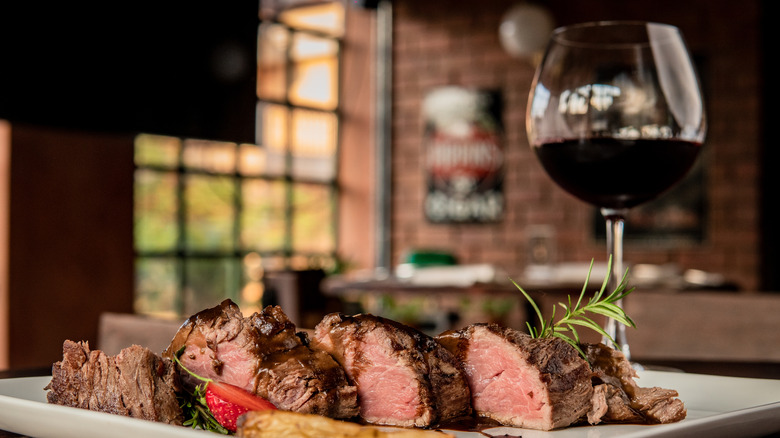 Glass of red wine next to sliced steak on white plate.