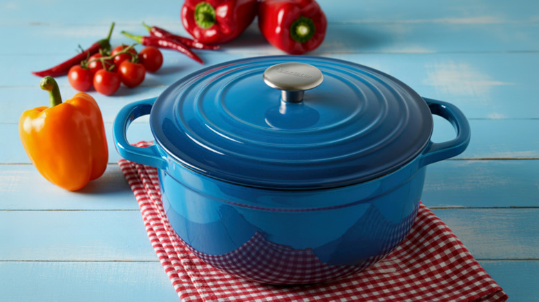 A vibrant blue enameled cast iron Dutch oven sits on a red and white checkered cloth, surrounded by fresh bell peppers and tomatoes.