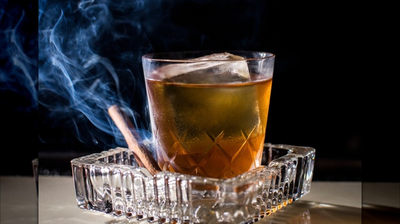 A cocktail on a crystal ash tray with a smoking cinnamon stick next to it