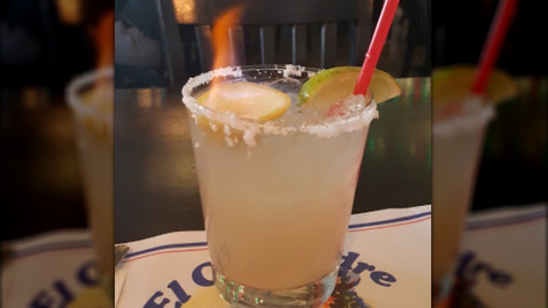 A margarita with a flaming lemon, lime garnish, salted rim, and red straw