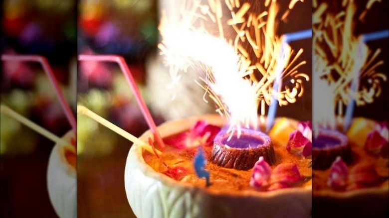 A close up of a drink in a shared bowl with multiple straws, cinnamon, tropical flowers, and sparks in the middle