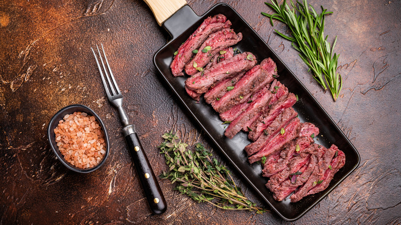 Sliced flank steak on a tray with salt and fresh herbs on the side.