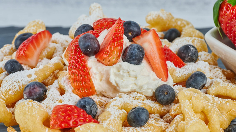 A deep-fried funnel cake covered in sliced strawberries, blueberries, and whipped cream