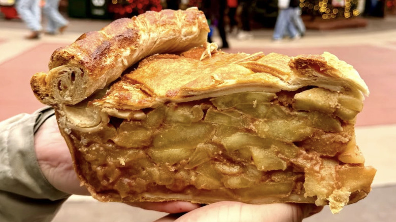 Two hands holding a huge slice of apple pie with a lattice crust and rolled edge.
